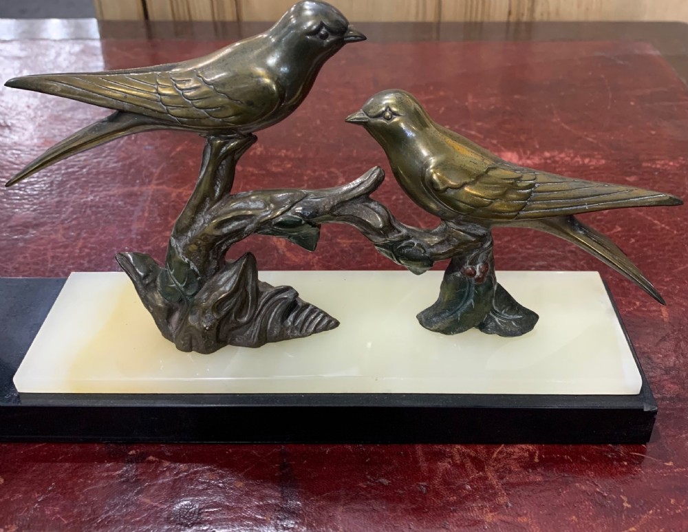 A French Art Deco mantel clock with a circular cream onyx timepiece mounted on a black marble base beside a metal bird sculpture. View II