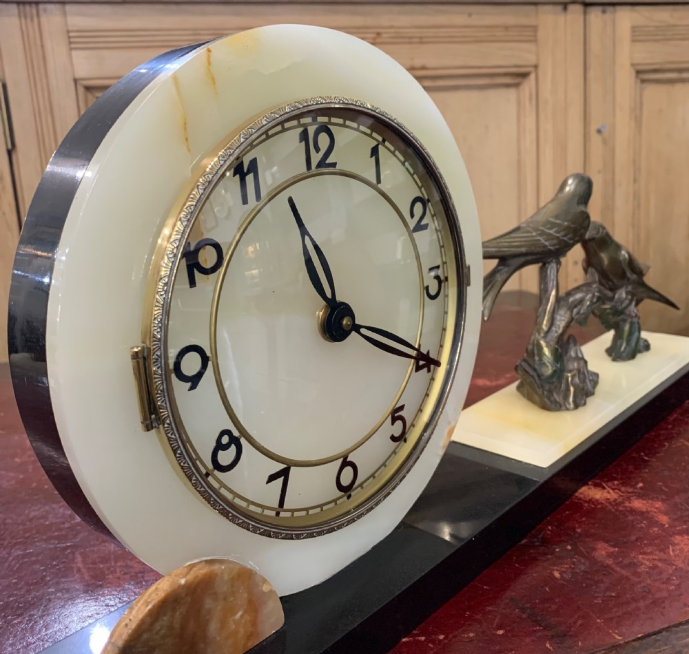 A French Art Deco mantel clock with a circular cream onyx timepiece mounted on a black marble base beside a metal bird sculpture. View VI