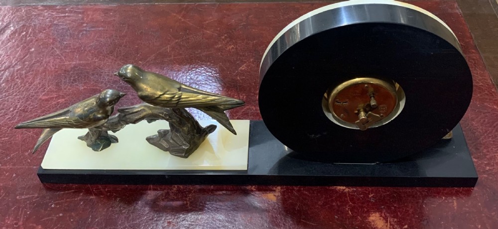 A French Art Deco mantel clock with a circular cream onyx timepiece mounted on a black marble base beside a metal bird sculpture. View VII