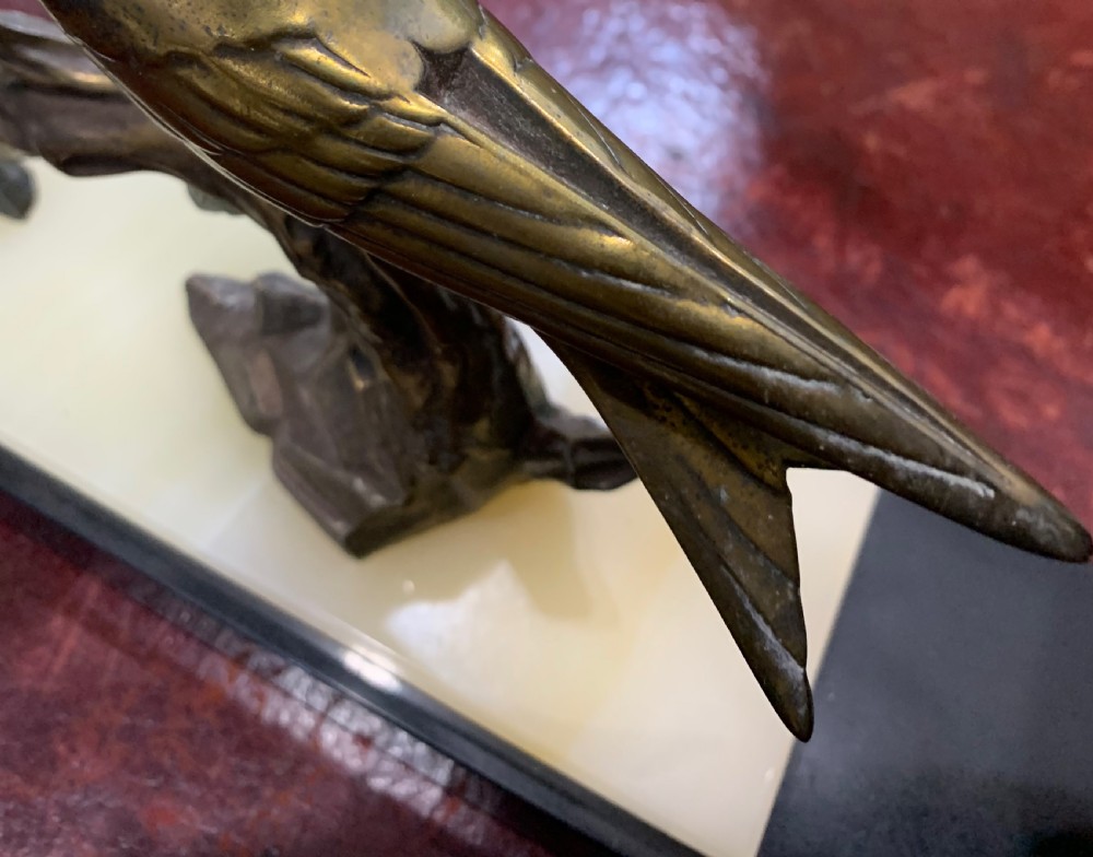 A French Art Deco mantel clock with a circular cream onyx timepiece mounted on a black marble base beside a metal bird sculpture. View IX