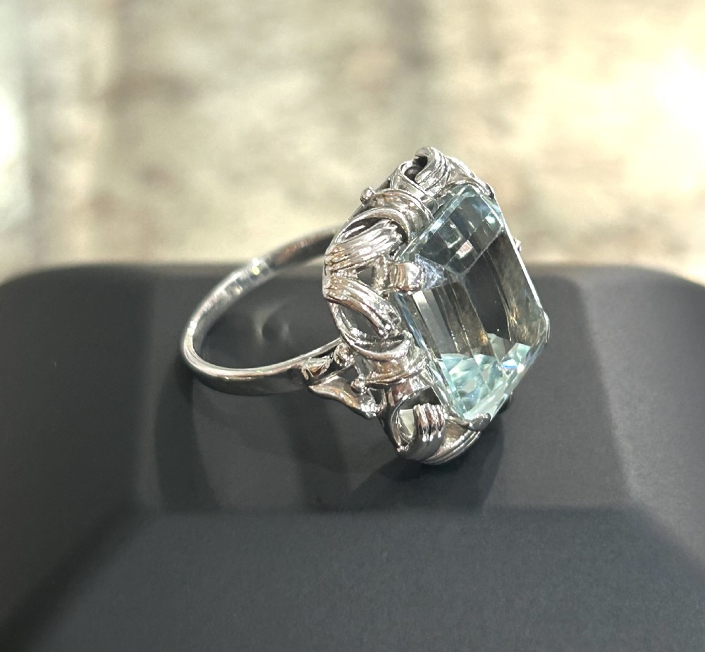 An emerald-cut light blue aquamarine gemstone set in a white gold claw mount inside a black presentation box. View VI