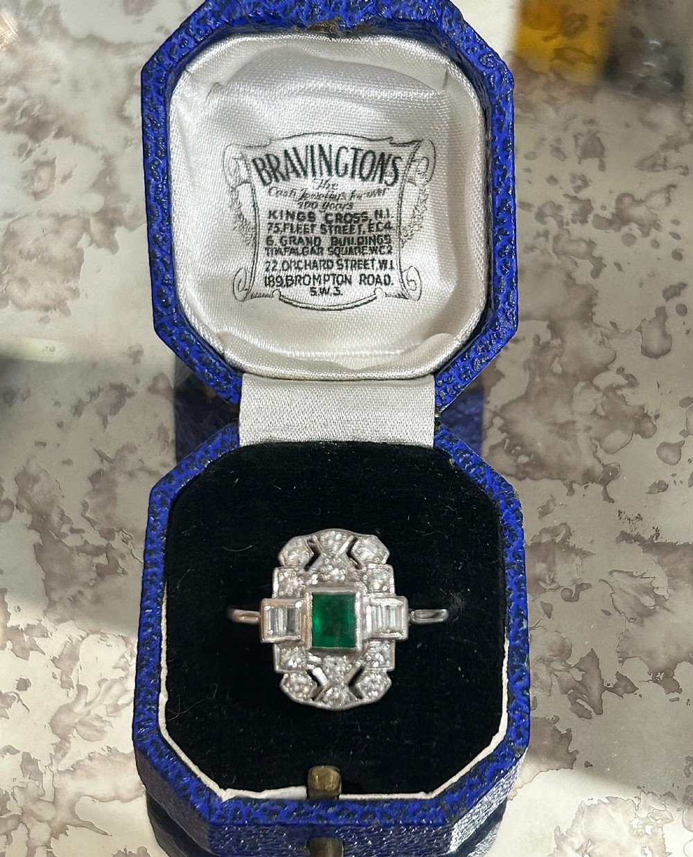 An Art Deco platinum cluster ring featuring a central emerald cut emerald framed by geometric baguette and brilliant cut diamonds, set inside a blue velvet presentation box.