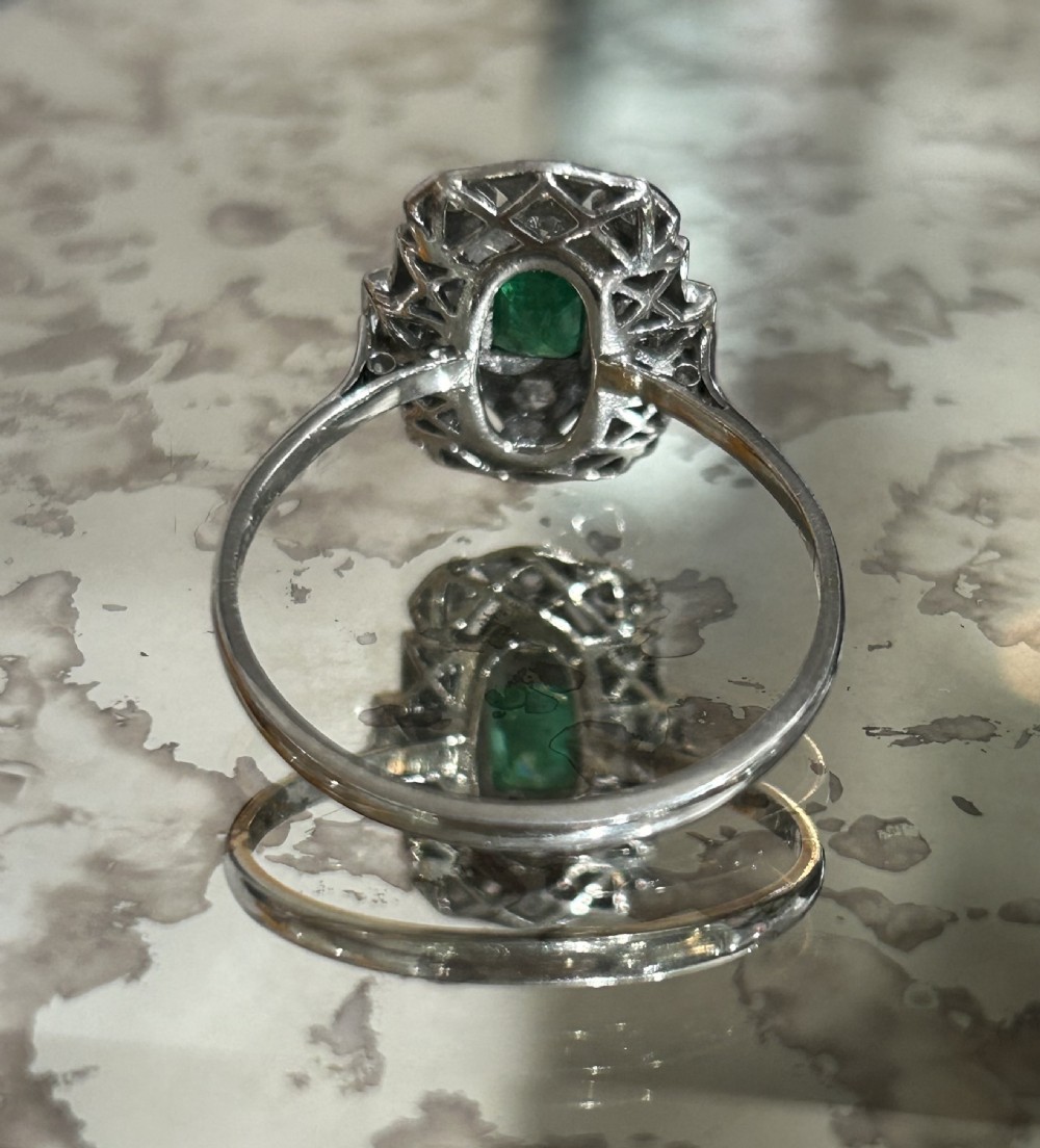 An Art Deco platinum cluster ring featuring a central emerald cut emerald framed by geometric baguette and brilliant cut diamonds, set inside a blue velvet presentation box. View III