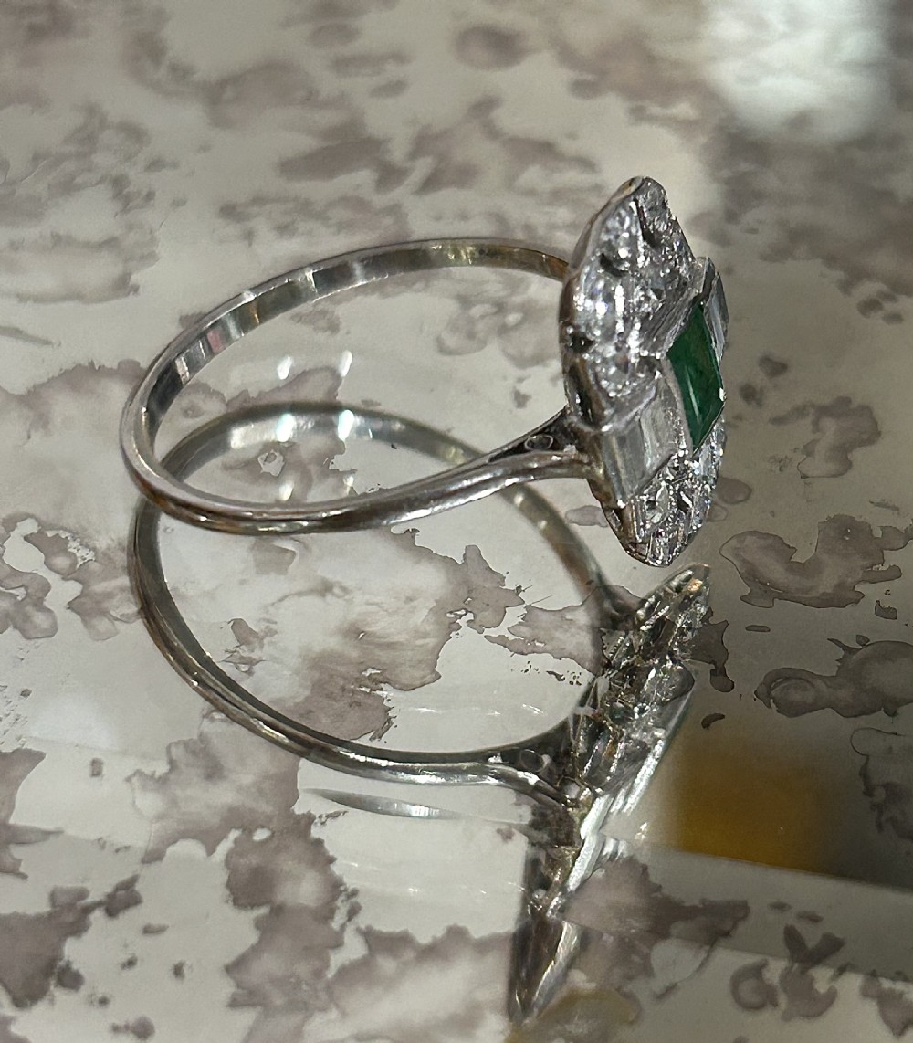 An Art Deco platinum cluster ring featuring a central emerald cut emerald framed by geometric baguette and brilliant cut diamonds, set inside a blue velvet presentation box. View V