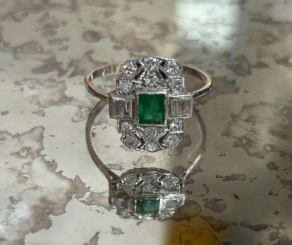 An Art Deco platinum cluster ring featuring a central emerald cut emerald framed by geometric baguette and brilliant cut diamonds, set inside a blue velvet presentation box. View VI