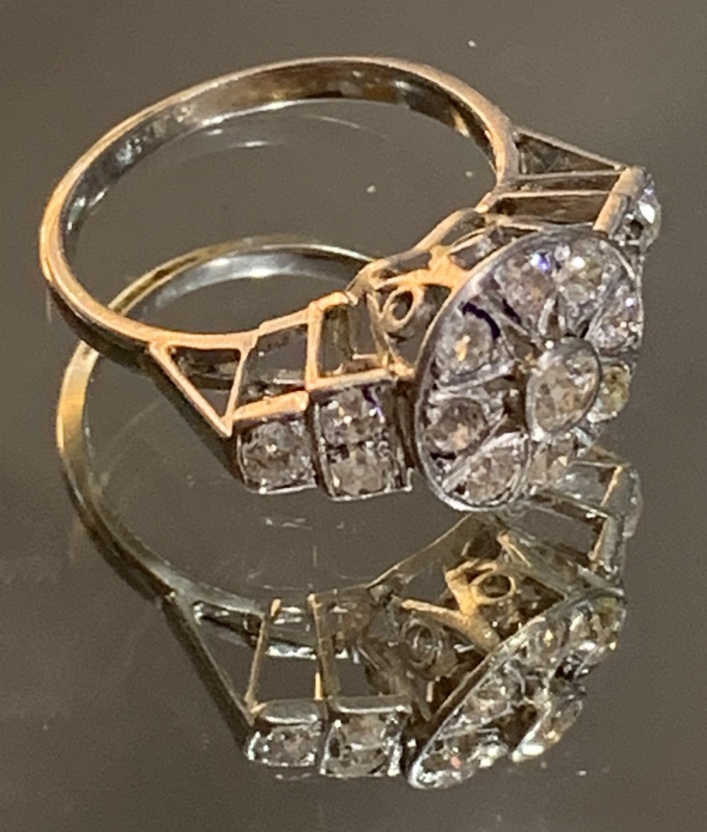 An Art Deco platinum cluster ring featuring a central round diamond framed by smaller diamonds in a geometric starburst setting with stepped shoulders. View VI