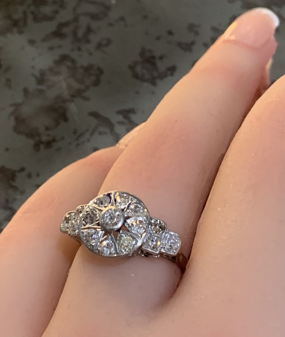 An Art Deco platinum cluster ring featuring a central round diamond framed by smaller diamonds in a geometric starburst setting with stepped shoulders. View VII