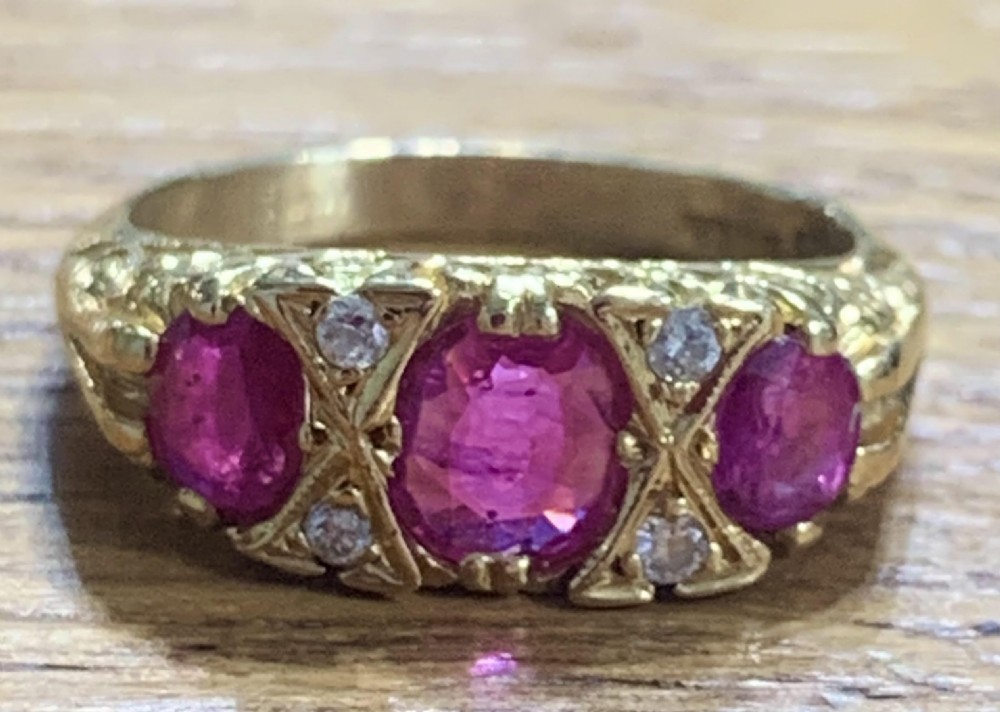An antique 18ct yellow gold trilogy ring featuring three oval cut rubies separated by small diamond accents set within a carved gold band.