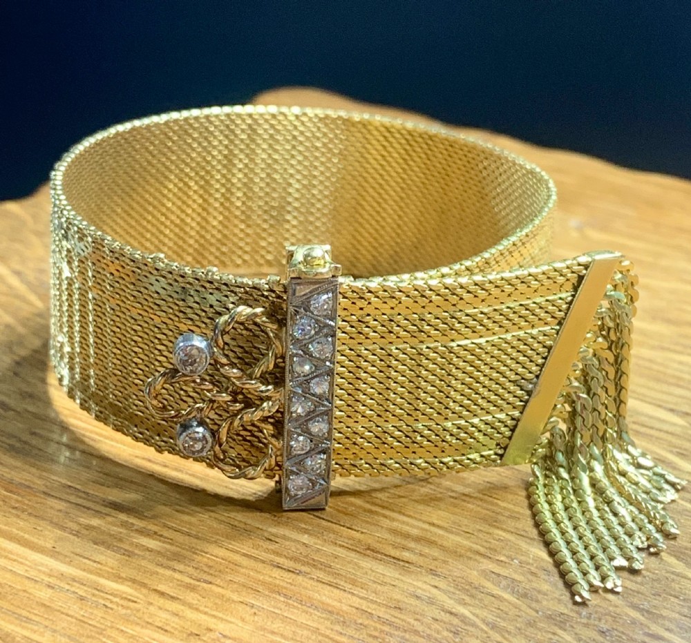 An Art Deco 18ct yellow gold mesh band bracelet featuring a diamond-set vertical bar, wire scrollwork, and a hanging gold tassel fringe.