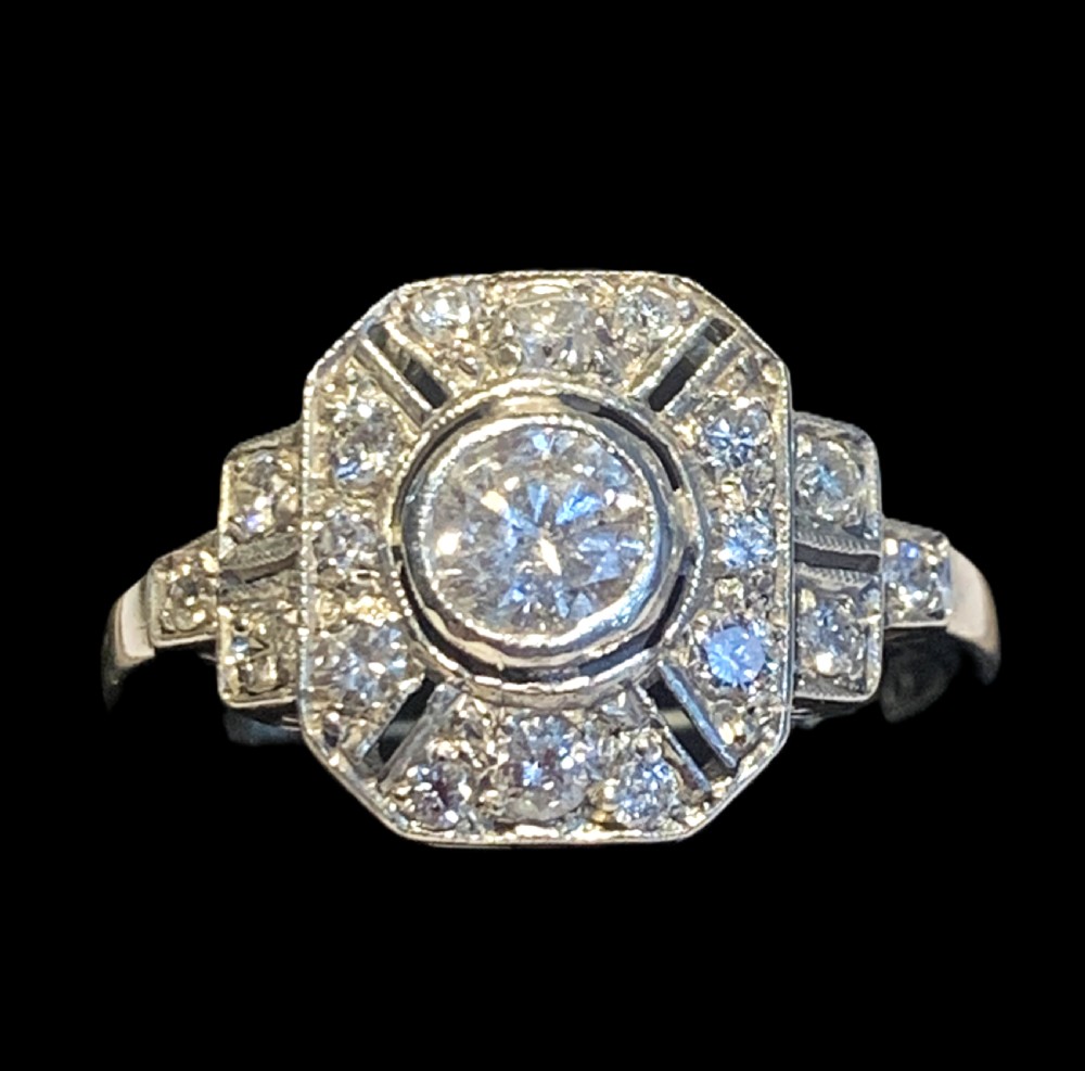An Art Deco 18ct white gold ring featuring a round central diamond enclosed within an octagonal halo setting with millegrain details.