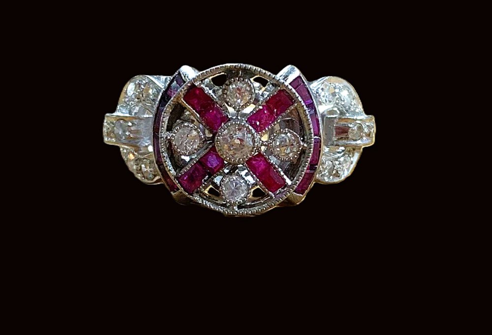 An Art Deco ring featuring a central round diamond encircled by geometric cross-shaped calibre-cut rubies and additional diamond accents set in white metal.