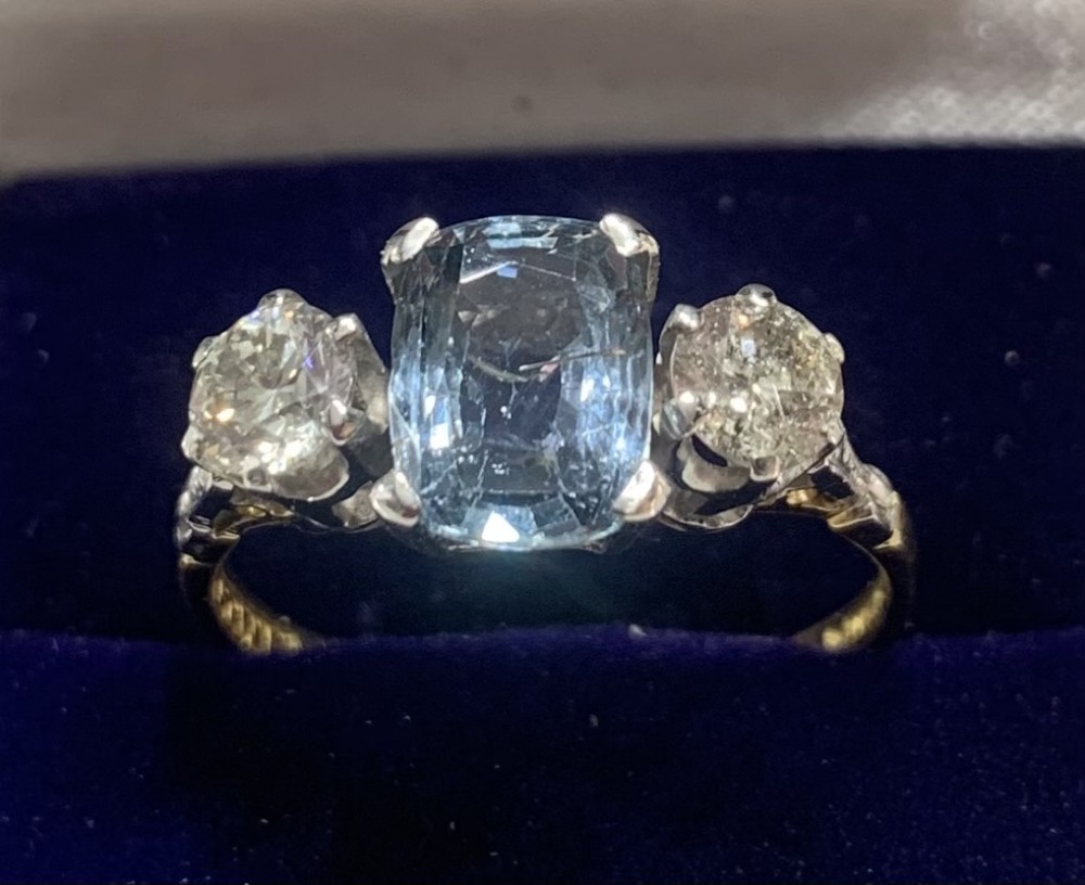 An Art Deco trilogy ring featuring a central cushion-cut light blue sapphire flanked by two brilliant-cut diamonds set in platinum claws on an eighteen-carat gold band.