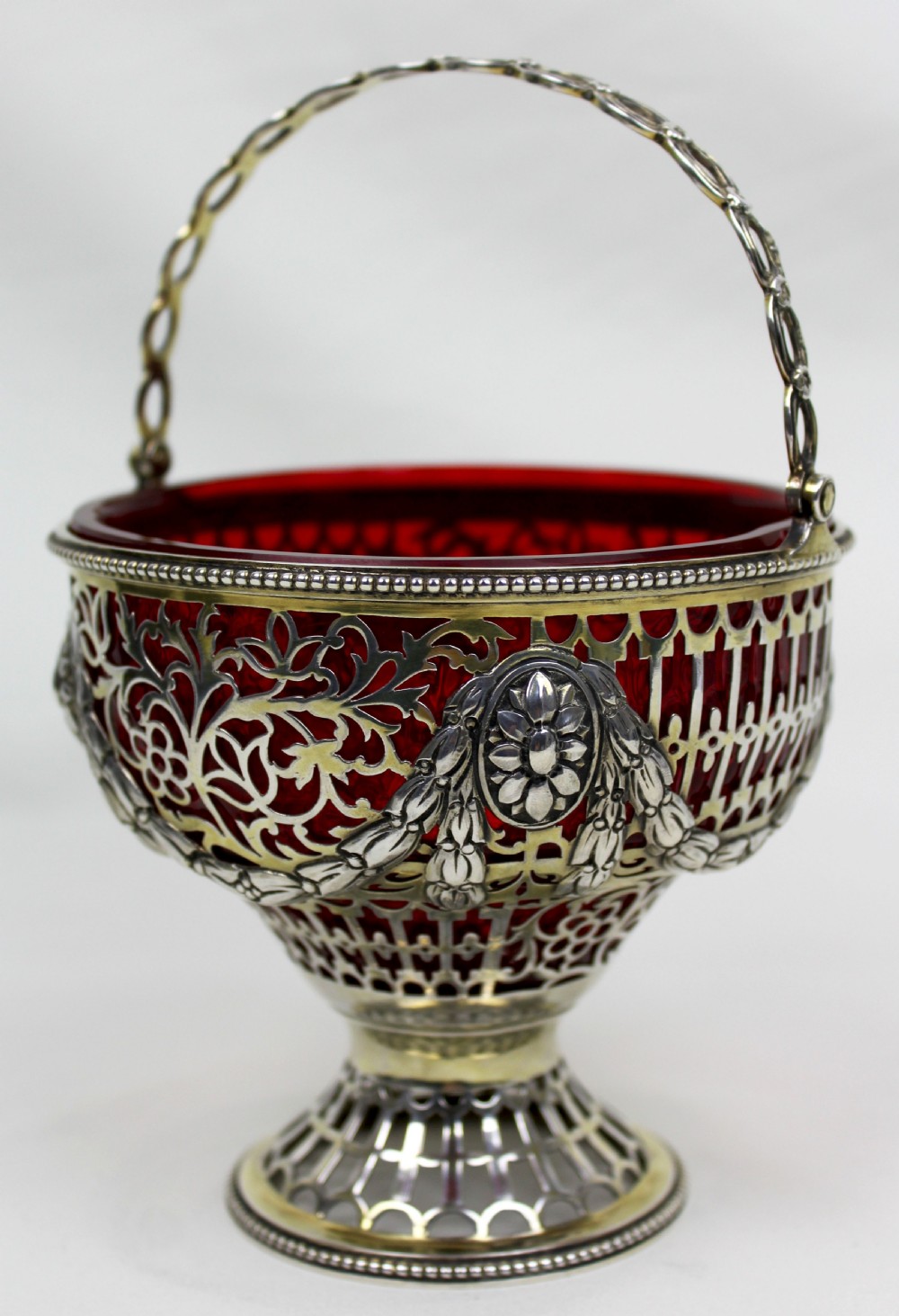 A Victorian silver-gilt sugar basket featuring elaborate pierced openwork, floral swags, a swing handle, and a removable ruby red glass liner.