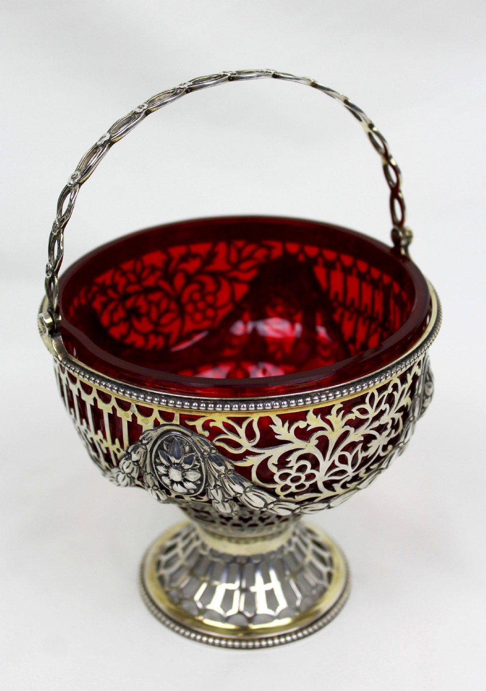 A Victorian silver-gilt sugar basket featuring elaborate pierced openwork, floral swags, a swing handle, and a removable ruby red glass liner. View III