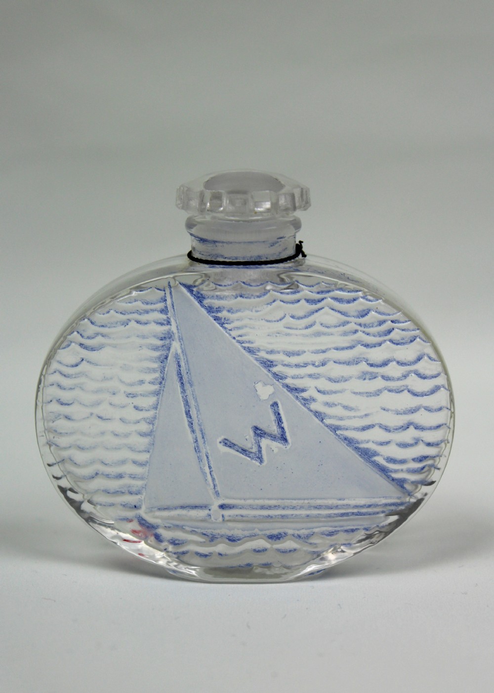 An oval clear glass scent bottle featuring a moulded sailboat on ocean waves accentuated with blue stain, topped with a faceted stopper.