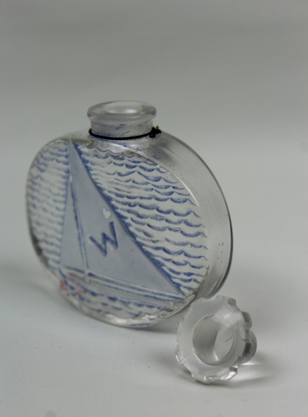 An oval clear glass scent bottle featuring a moulded sailboat on ocean waves accentuated with blue stain, topped with a faceted stopper. View III