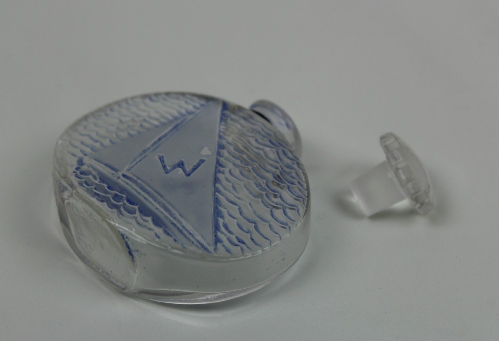 An oval clear glass scent bottle featuring a moulded sailboat on ocean waves accentuated with blue stain, topped with a faceted stopper. View IV