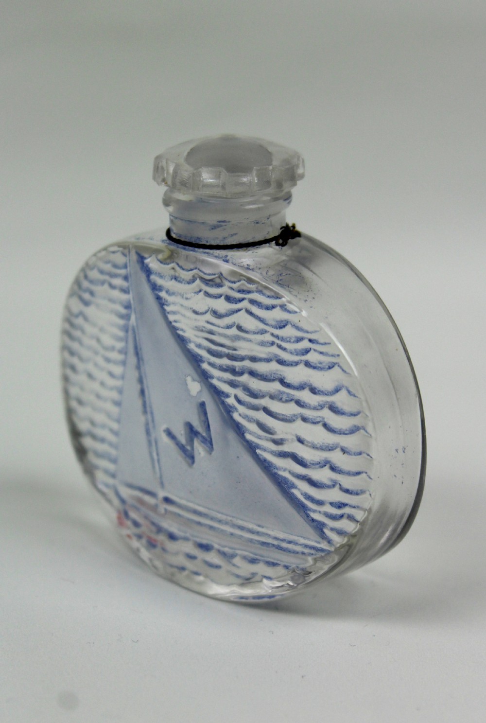 An oval clear glass scent bottle featuring a moulded sailboat on ocean waves accentuated with blue stain, topped with a faceted stopper. View V