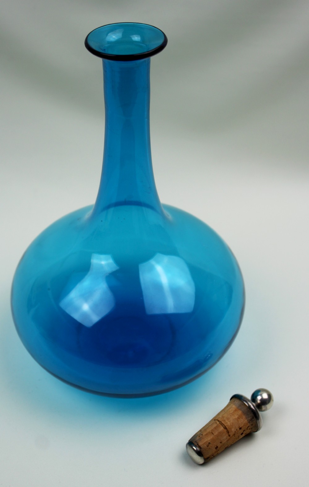 A vibrant blue hand-blown glass mell decanter with a rounded base, slender neck, and a cork stopper topped with a silver spherical finial. View II