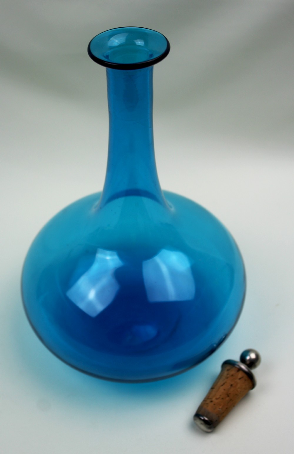 A vibrant blue hand-blown glass mell decanter with a rounded base, slender neck, and a cork stopper topped with a silver spherical finial. View IV