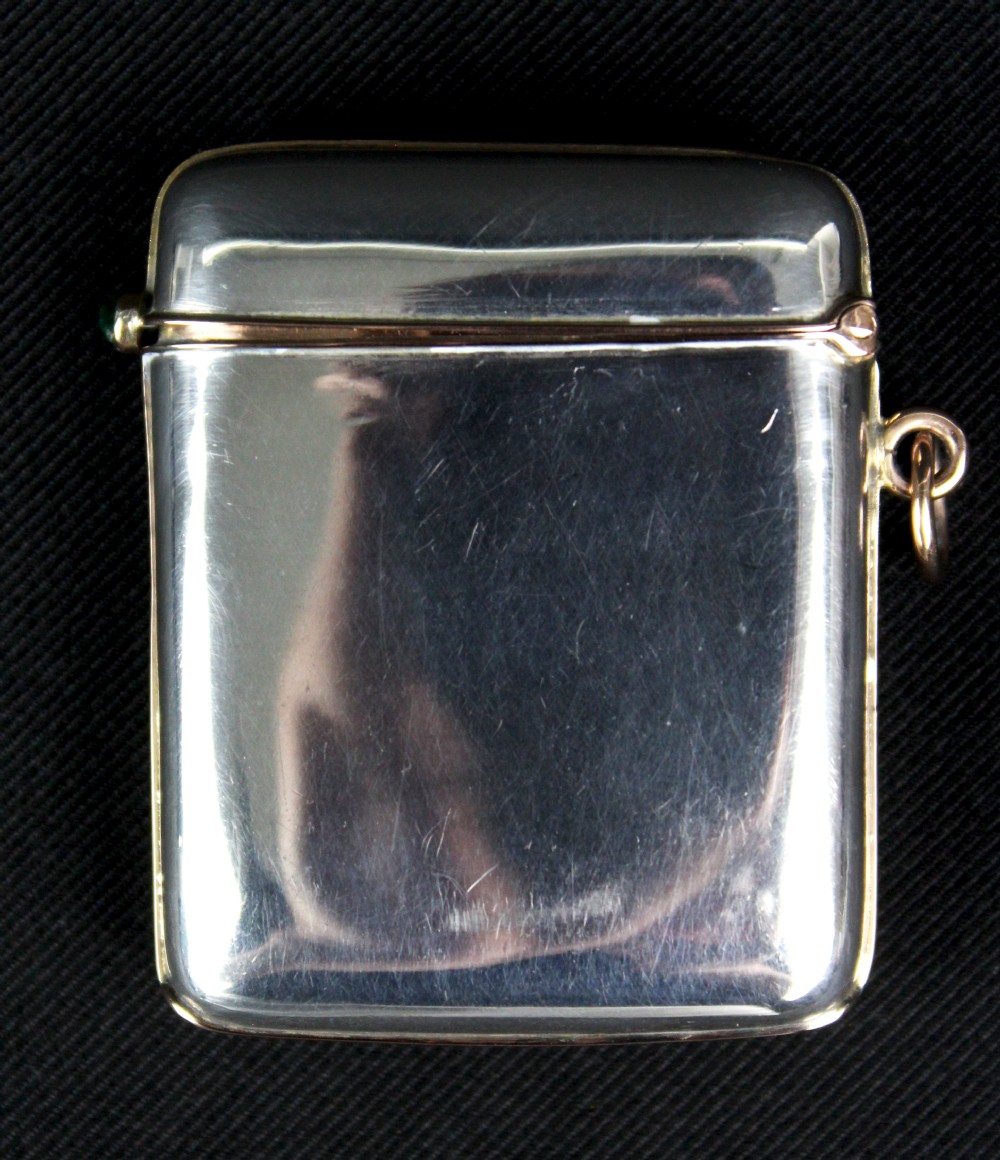 An antique rectangular sterling silver vesta case featuring gold trim, a side attachment loop, and a shamrock motif set with green cabochon gemstones and diamonds. View V