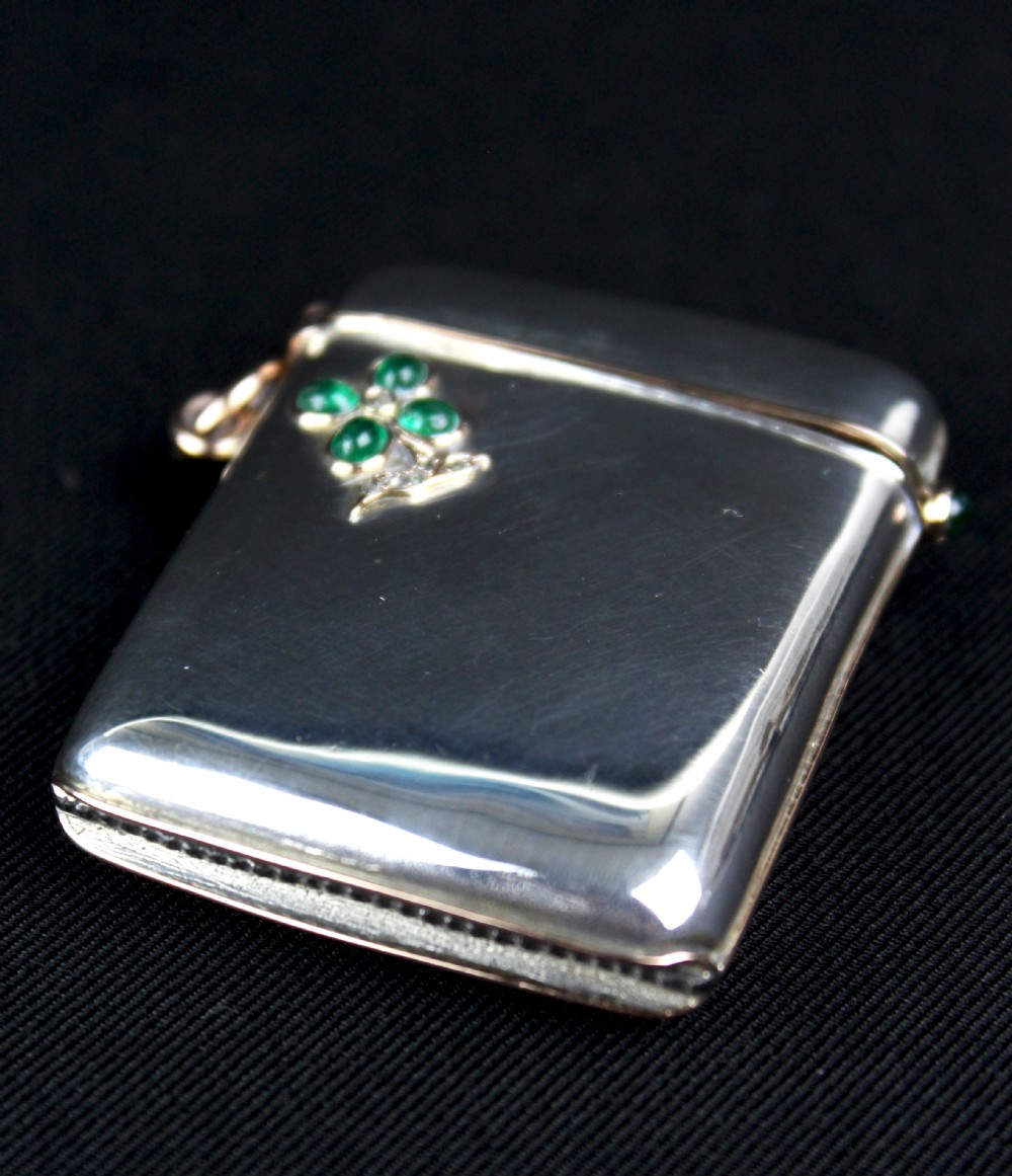 An antique rectangular sterling silver vesta case featuring gold trim, a side attachment loop, and a shamrock motif set with green cabochon gemstones and diamonds. View VI