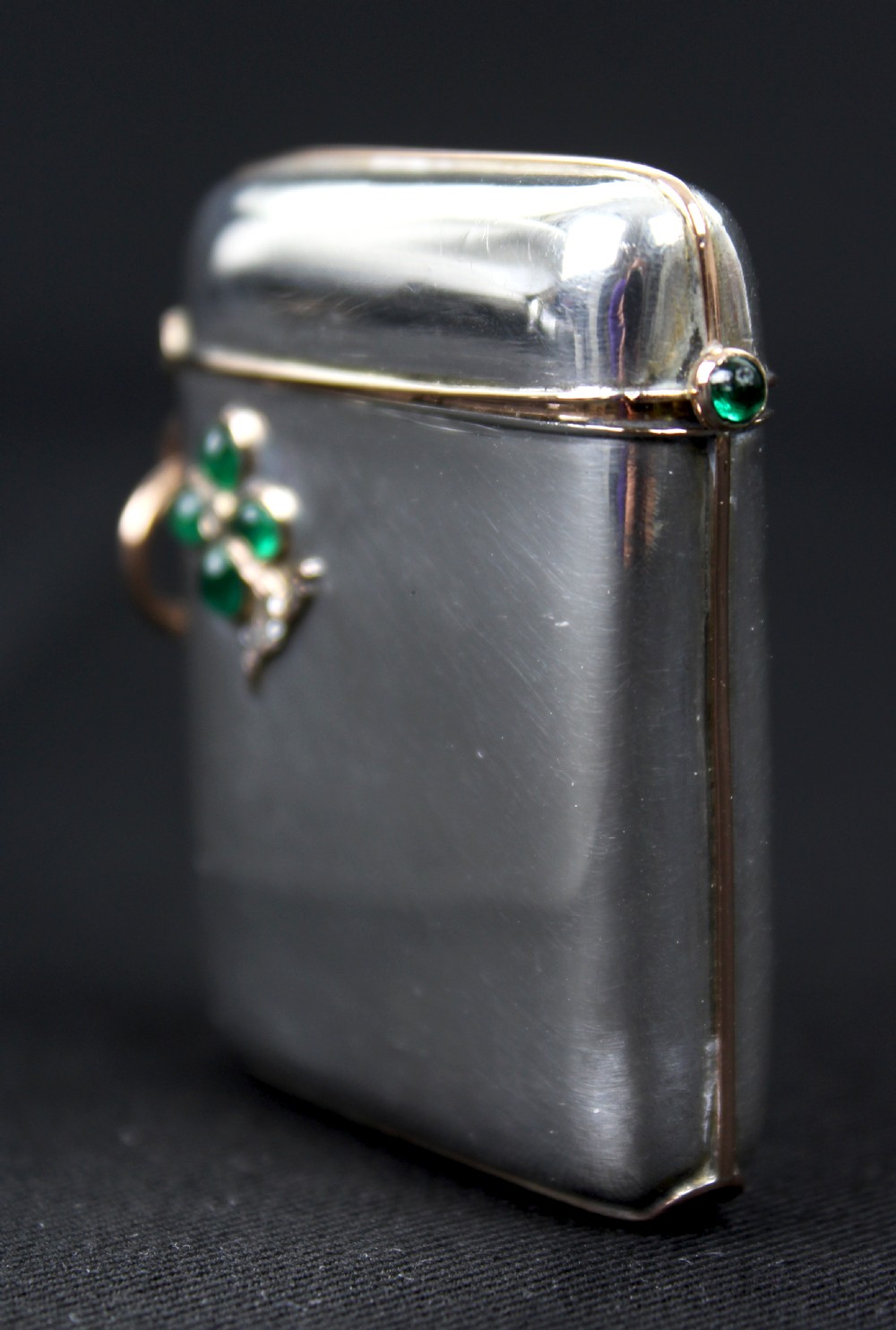 An antique rectangular sterling silver vesta case featuring gold trim, a side attachment loop, and a shamrock motif set with green cabochon gemstones and diamonds. View VII