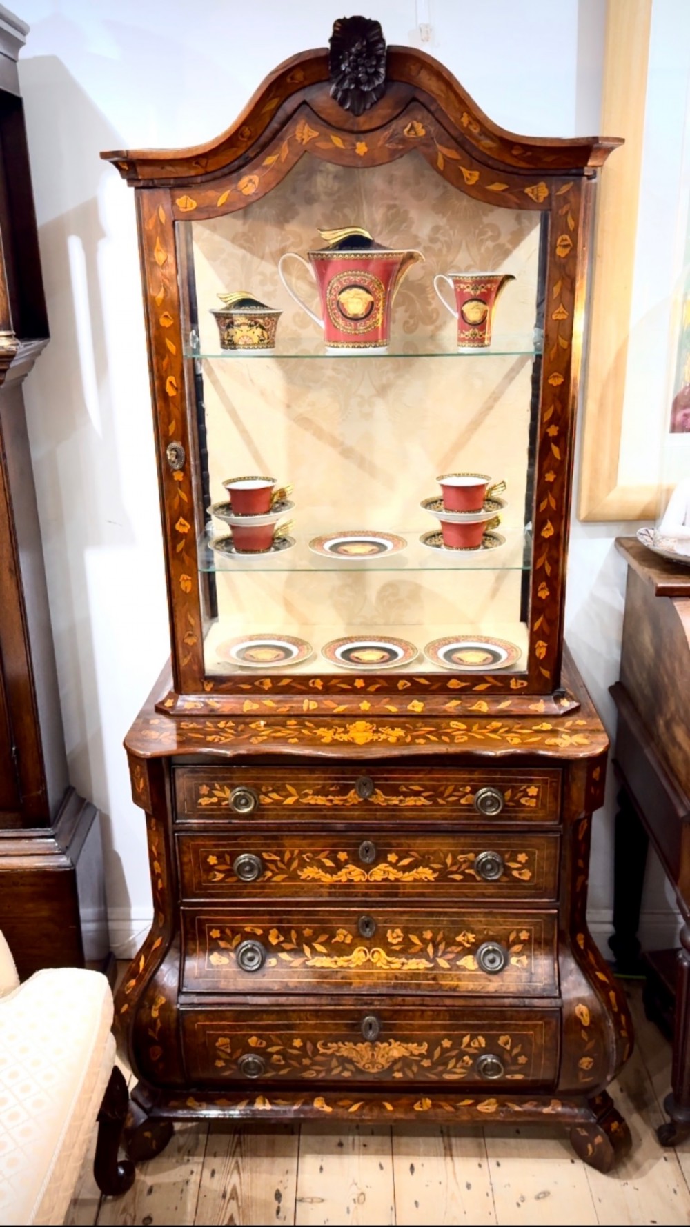 An antique Dutch walnut display cabinet featuring intricate floral wood marquetry, an arched glazed top, and a base with four drawers.