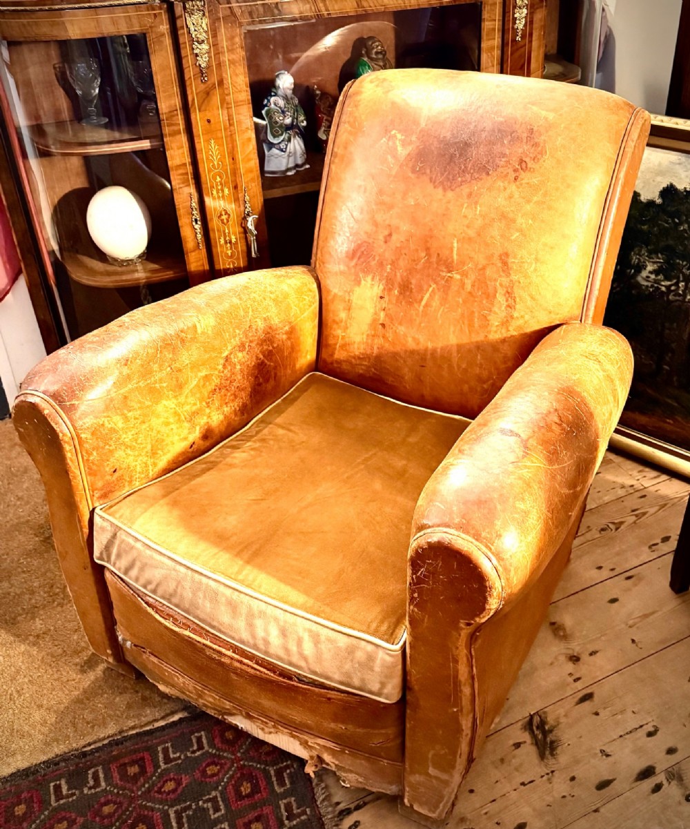 An Art Deco tan leather club chair featuring rolled armrests, distressed leather patina, and a fitted velvet seat cushion.