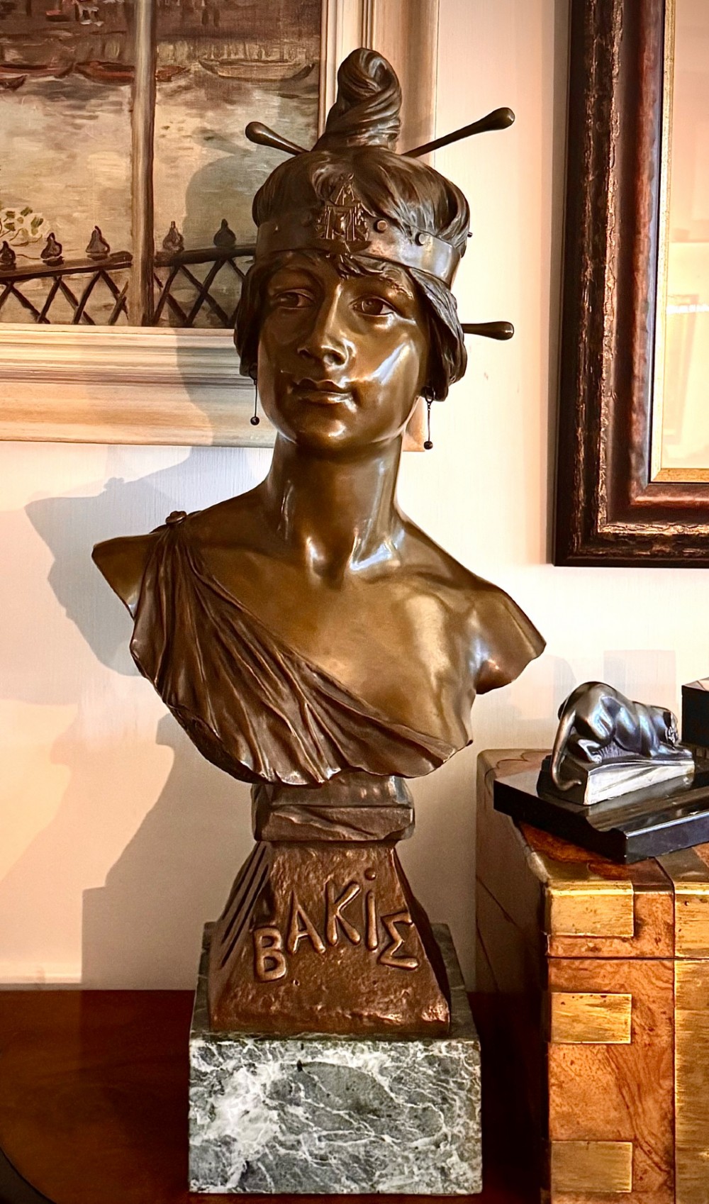 A patinated bronze bust of a female figure wearing a decorative headband and draped attire, mounted on an inscribed bronze pedestal atop a grey marble plinth.