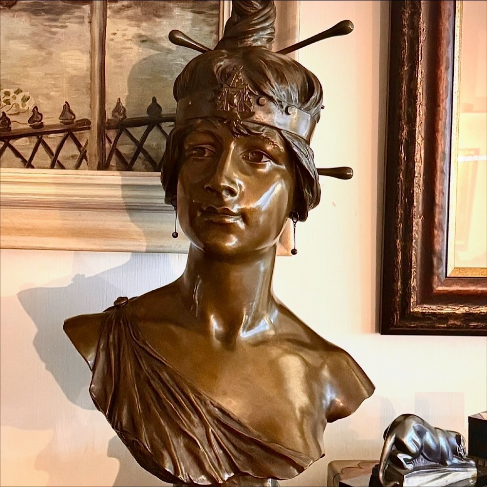 A patinated bronze bust of a female figure wearing a decorative headband and draped attire, mounted on an inscribed bronze pedestal atop a grey marble plinth. View II