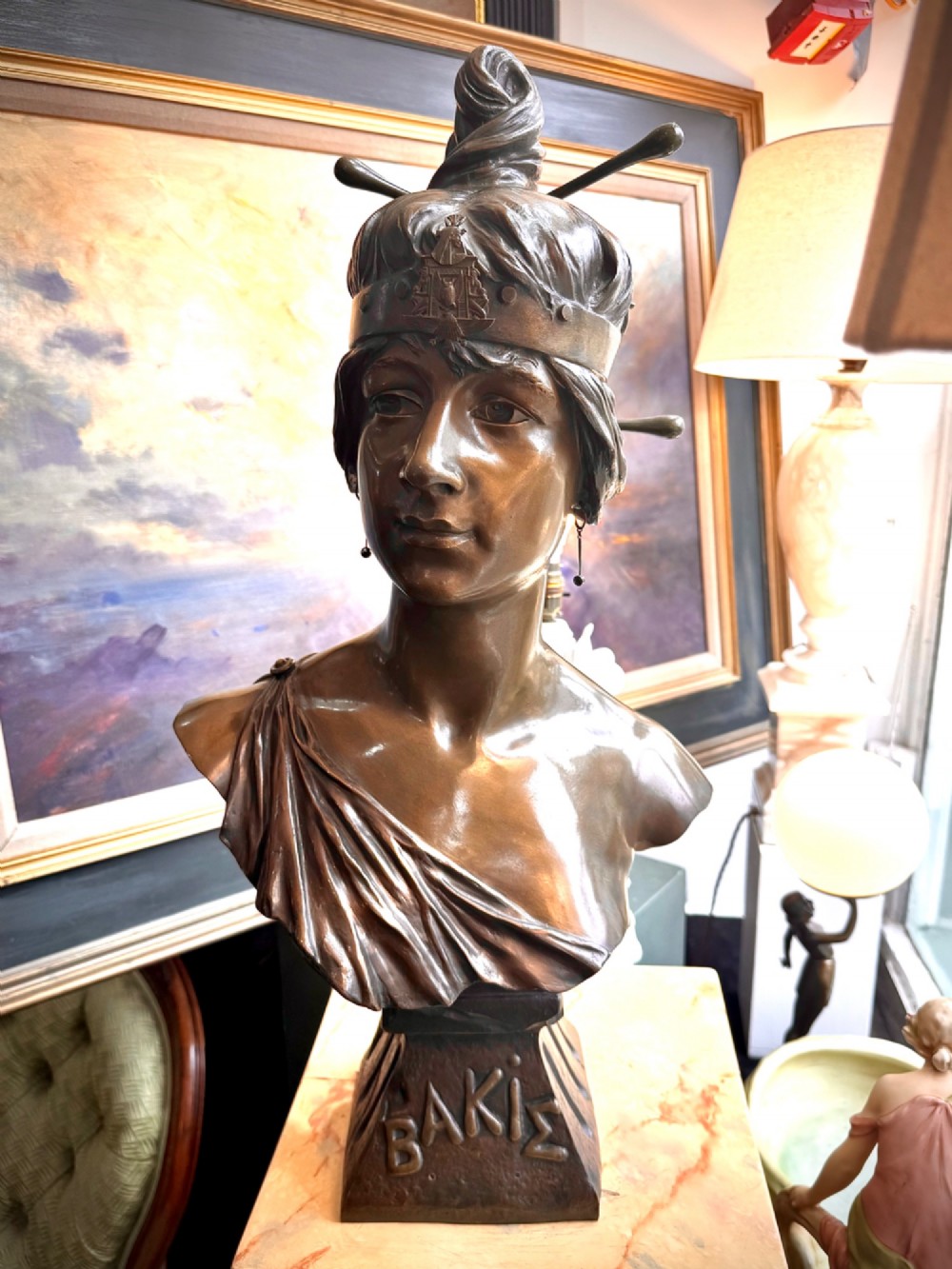 A patinated bronze bust of a female figure wearing a decorative headband and draped attire, mounted on an inscribed bronze pedestal atop a grey marble plinth. View V