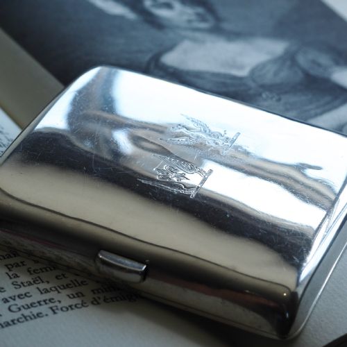 fig 5. A polished Victorian sterling silver cigarette case featuring an engraved heraldic crest and curved profile resting on an open book.
