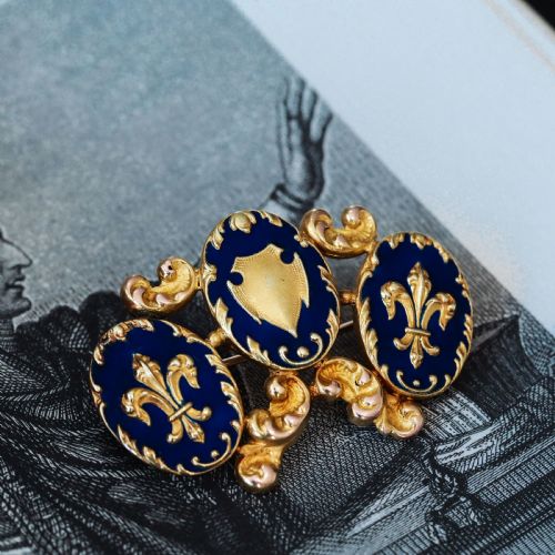 fig 2. A 19th-century 18ct gold brooch featuring three dark blue enamel oval cartouches decorated with gold heraldic motifs and rococo scrollwork.