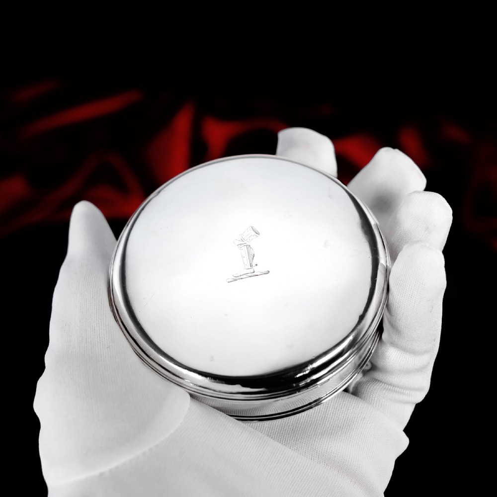 A circular Georgian sterling silver tobacco box featuring a polished flat cover hand-engraved with a crest.