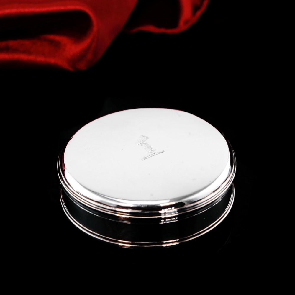 A circular Georgian sterling silver tobacco box featuring a polished flat cover hand-engraved with a crest. View VI
