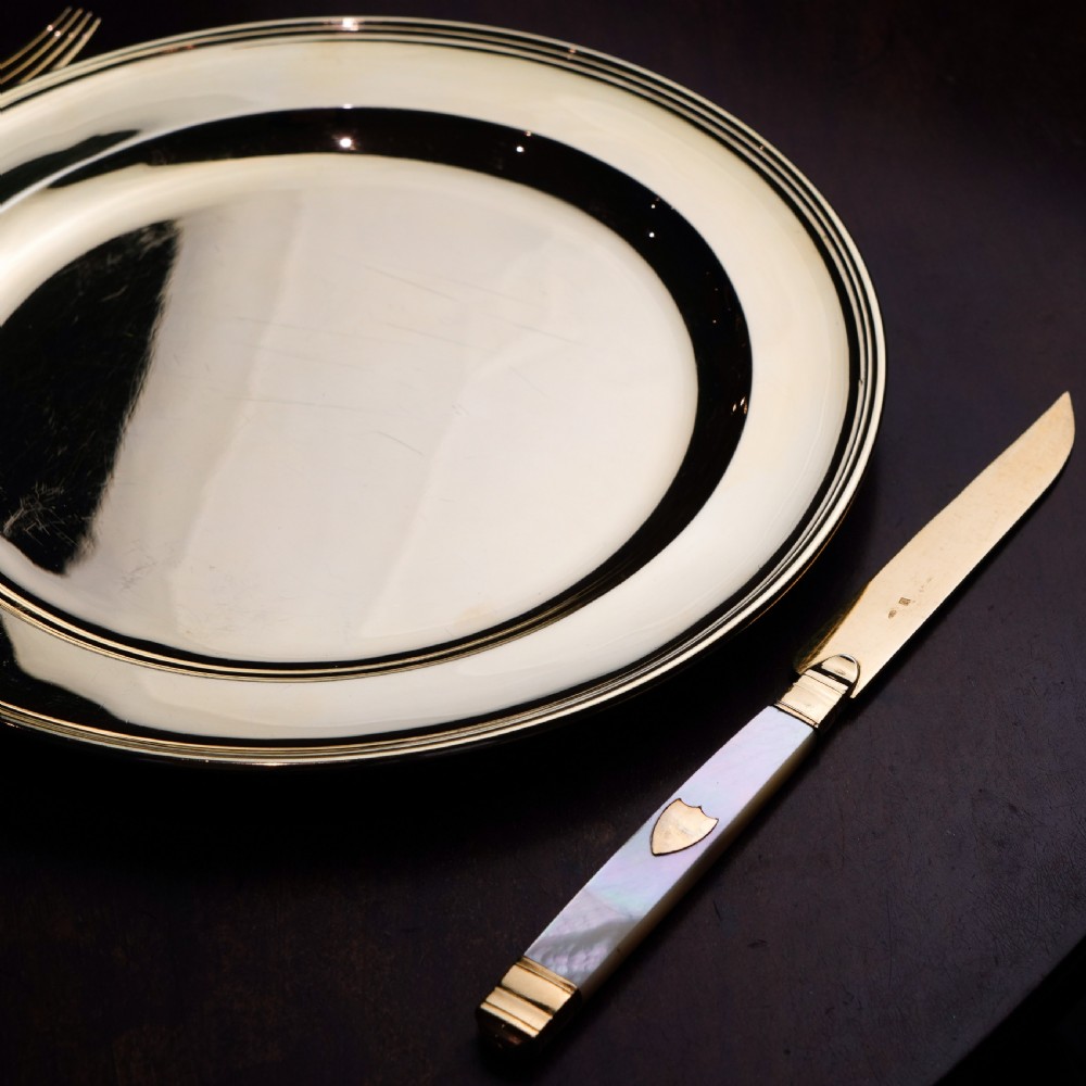 A set of six 19th-century fruit knives featuring silver-gilt blades and mother-of-pearl handles inlaid with small shield motifs. View X