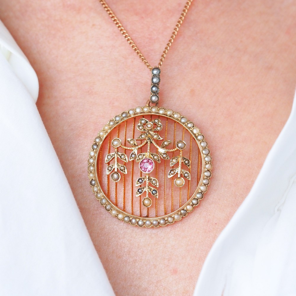 An Edwardian circular gold pendant featuring a halo of seed pearls framing an inner wire grid with a pearl-set floral garland and a central round pink tourmaline.