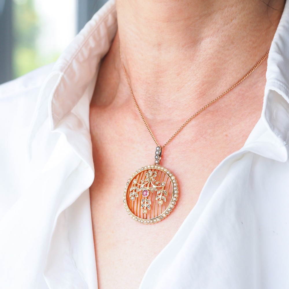 An Edwardian circular gold pendant featuring a halo of seed pearls framing an inner wire grid with a pearl-set floral garland and a central round pink tourmaline. View II