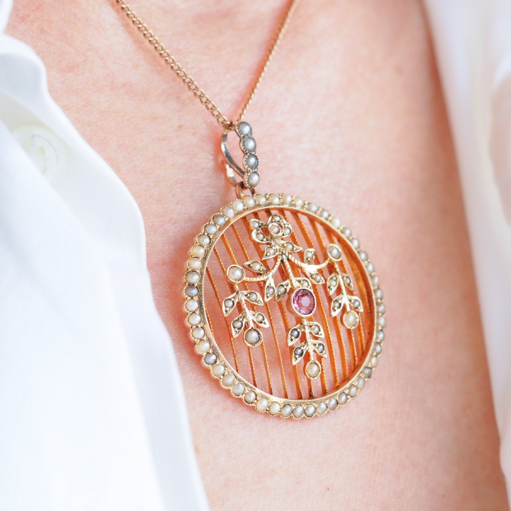 An Edwardian circular gold pendant featuring a halo of seed pearls framing an inner wire grid with a pearl-set floral garland and a central round pink tourmaline. View III
