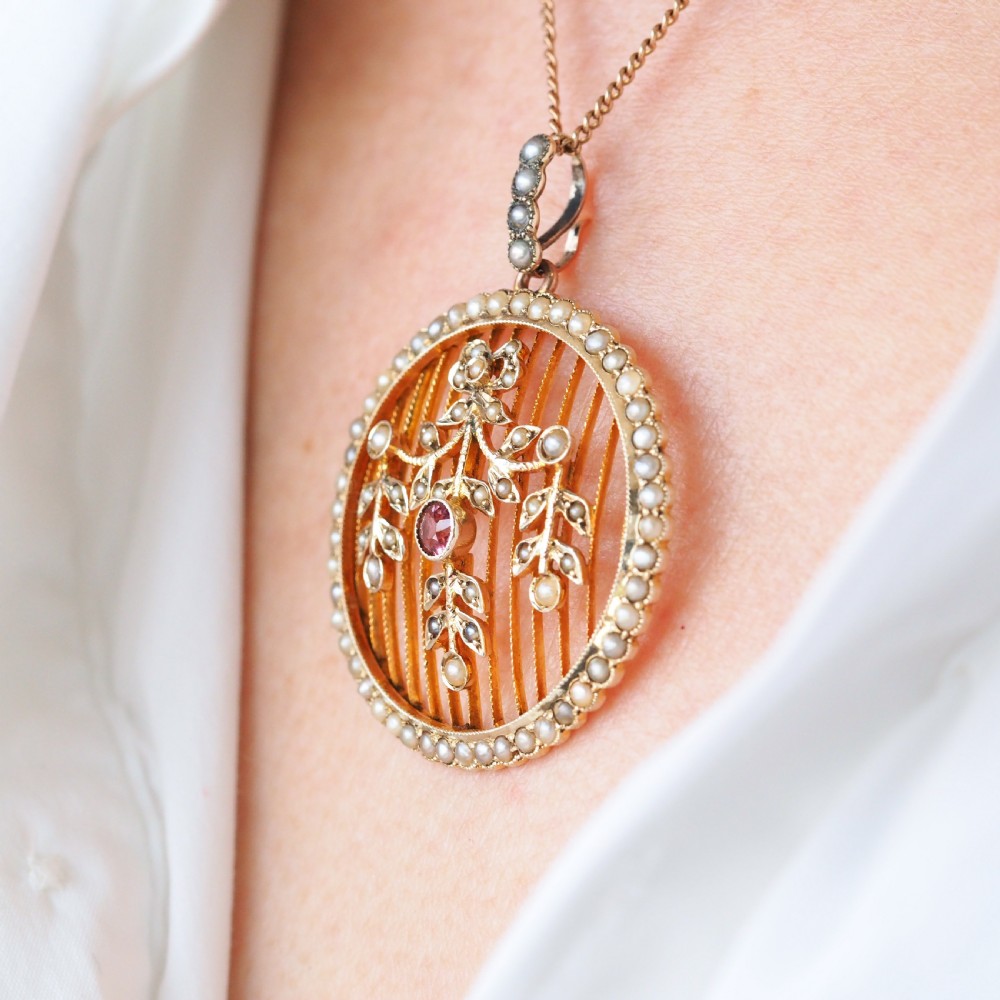 An Edwardian circular gold pendant featuring a halo of seed pearls framing an inner wire grid with a pearl-set floral garland and a central round pink tourmaline. View IV