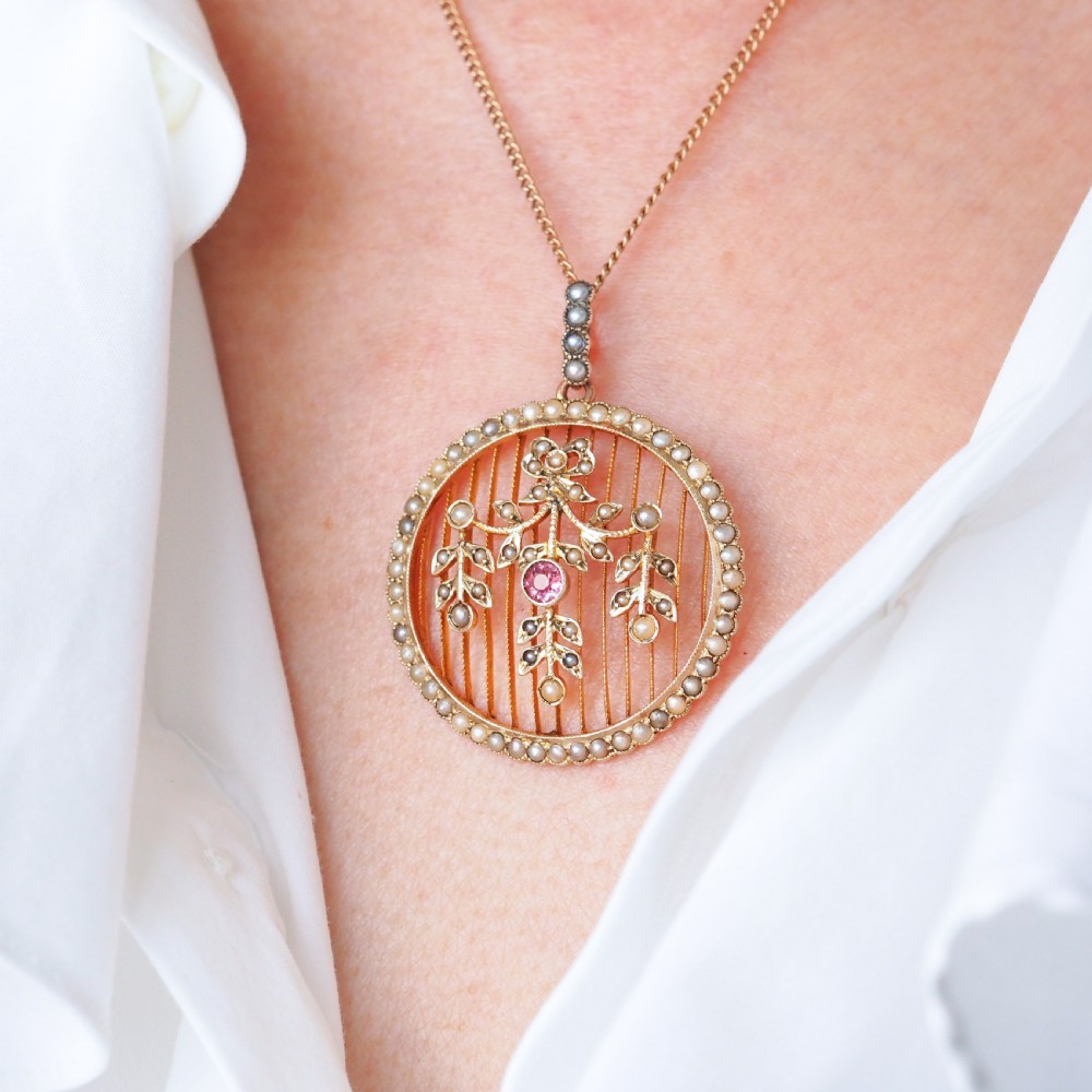 An Edwardian circular gold pendant featuring a halo of seed pearls framing an inner wire grid with a pearl-set floral garland and a central round pink tourmaline. View V