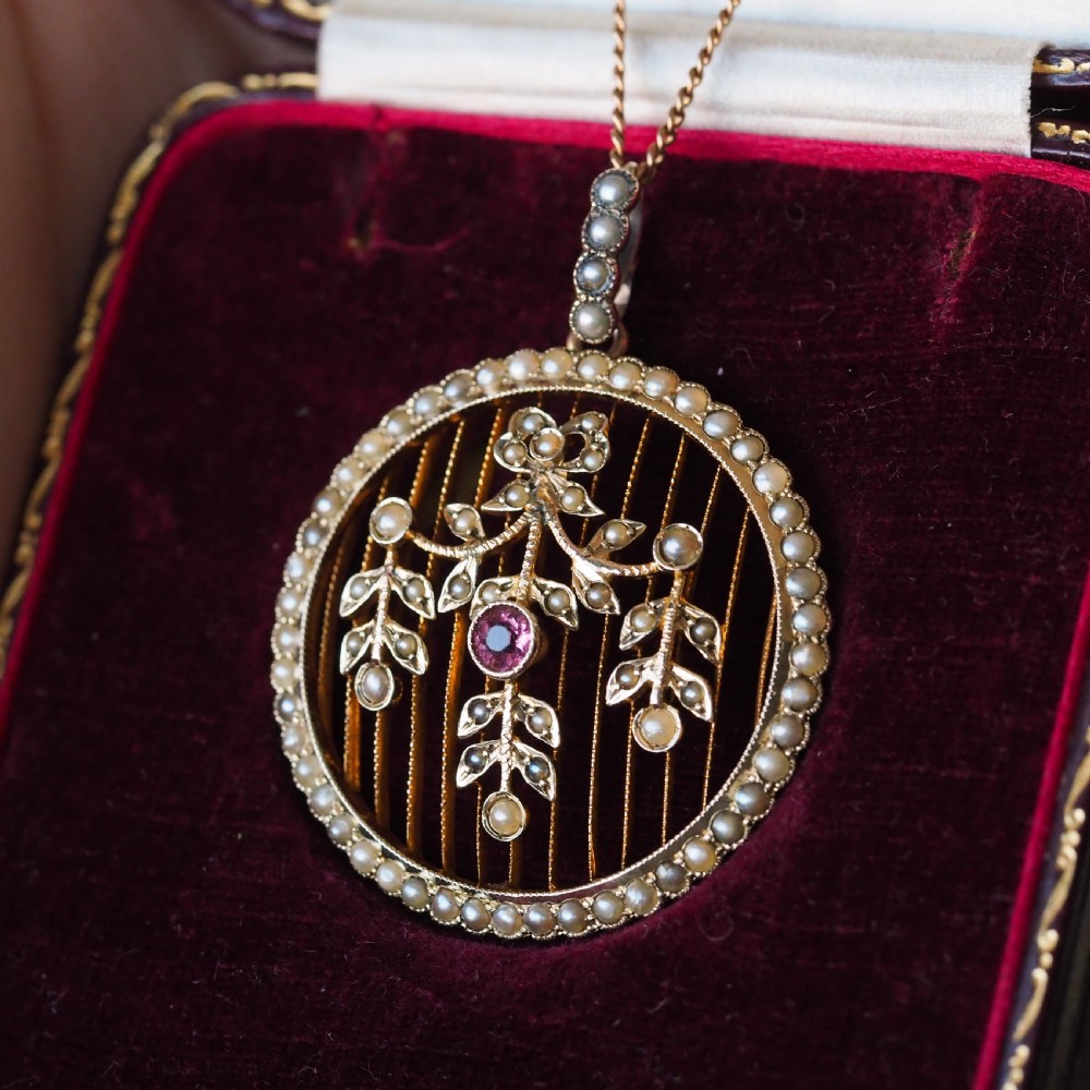 An Edwardian circular gold pendant featuring a halo of seed pearls framing an inner wire grid with a pearl-set floral garland and a central round pink tourmaline. View VI