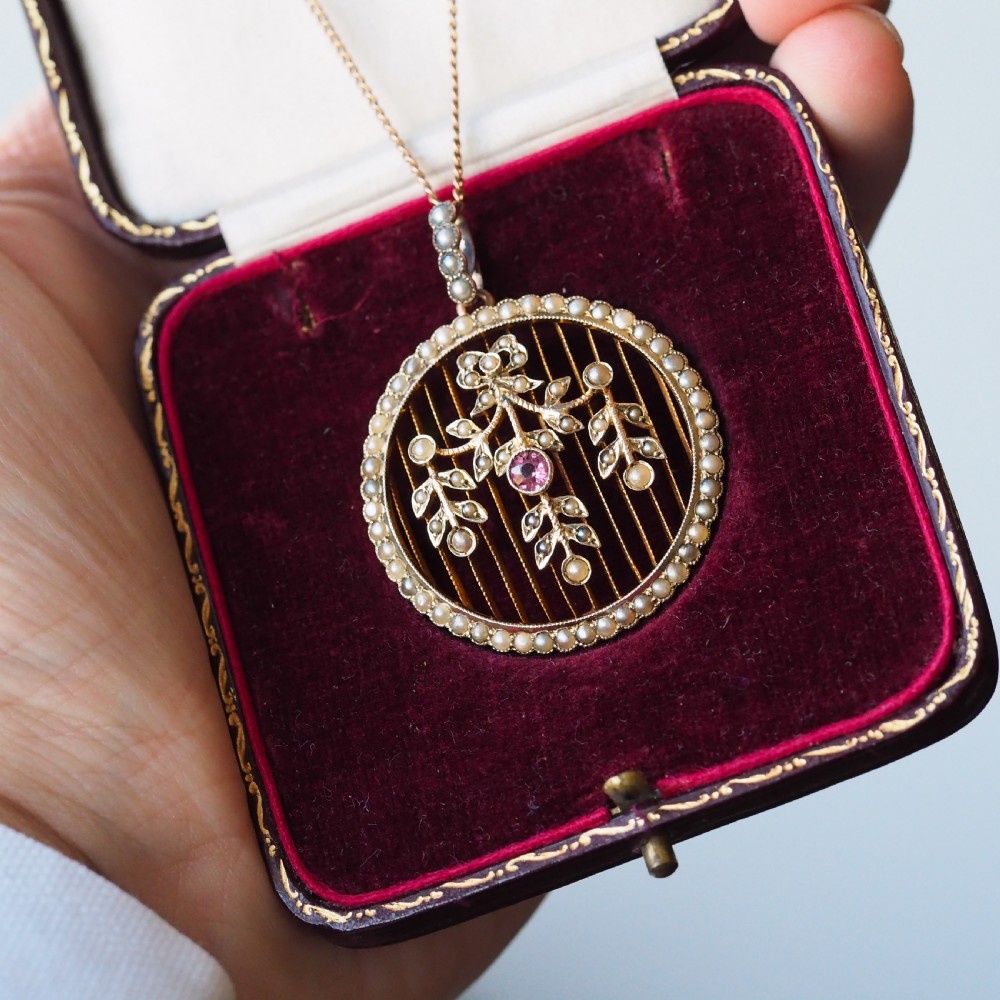 An Edwardian circular gold pendant featuring a halo of seed pearls framing an inner wire grid with a pearl-set floral garland and a central round pink tourmaline. View VII