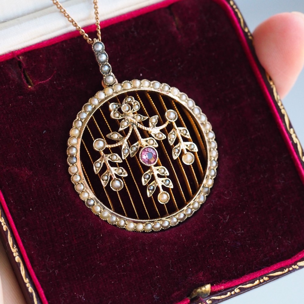 An Edwardian circular gold pendant featuring a halo of seed pearls framing an inner wire grid with a pearl-set floral garland and a central round pink tourmaline. View VIII