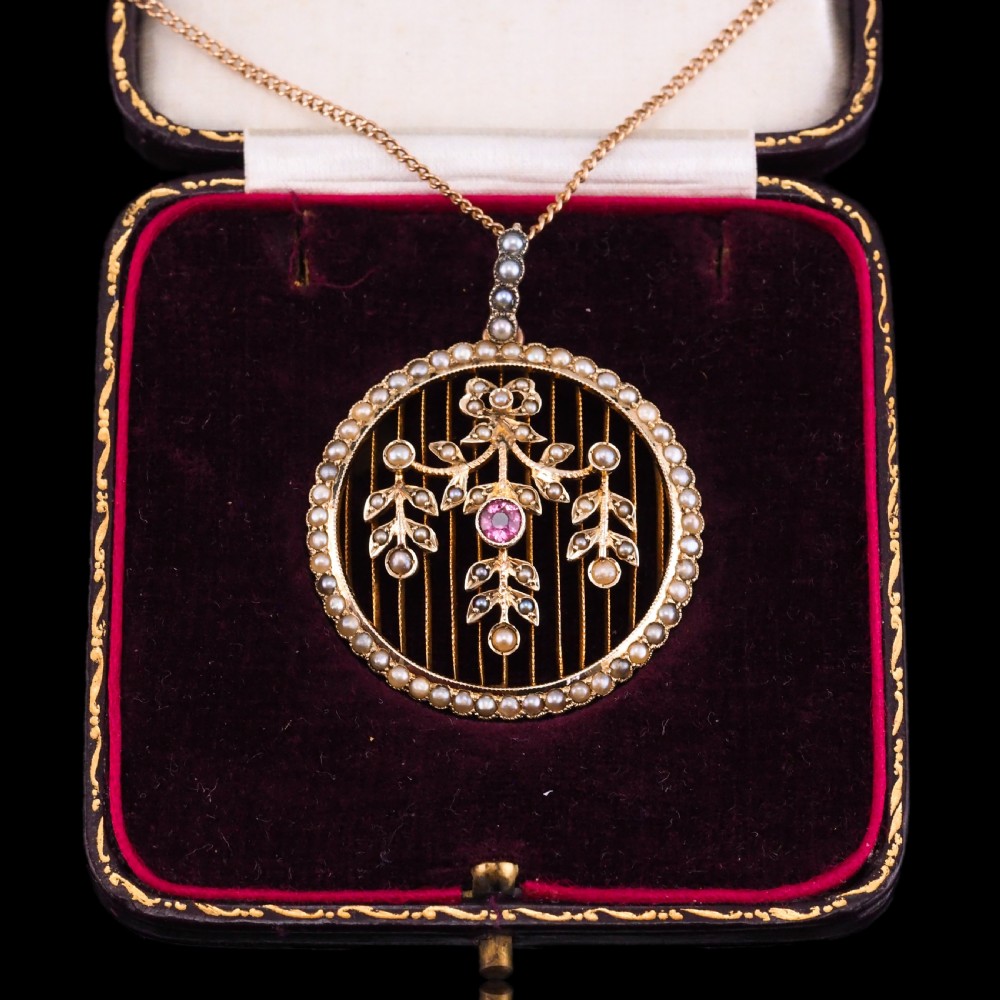 An Edwardian circular gold pendant featuring a halo of seed pearls framing an inner wire grid with a pearl-set floral garland and a central round pink tourmaline. View IX