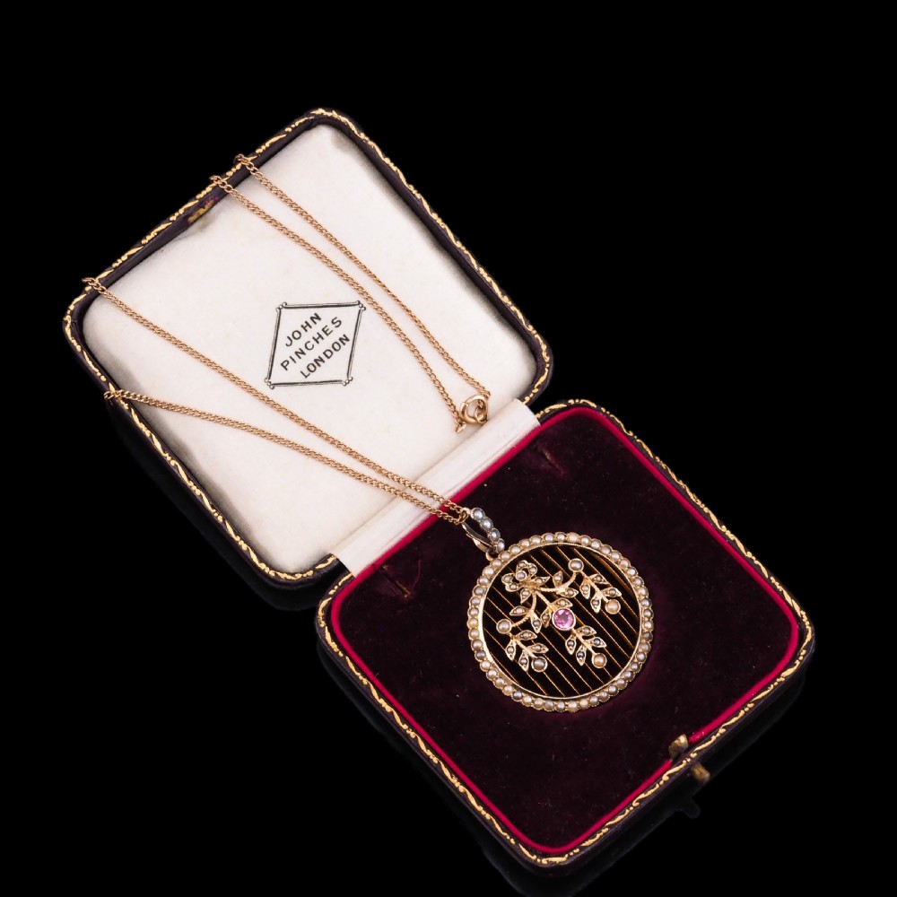 An Edwardian circular gold pendant featuring a halo of seed pearls framing an inner wire grid with a pearl-set floral garland and a central round pink tourmaline. View X