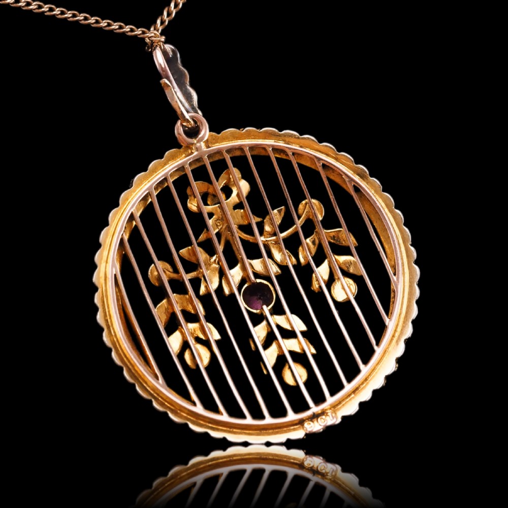 An Edwardian circular gold pendant featuring a halo of seed pearls framing an inner wire grid with a pearl-set floral garland and a central round pink tourmaline. View XII