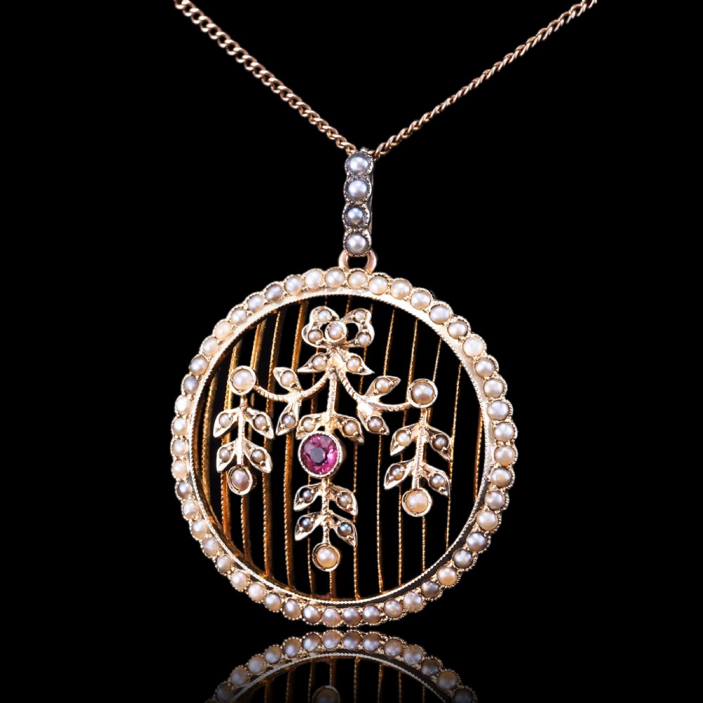 An Edwardian circular gold pendant featuring a halo of seed pearls framing an inner wire grid with a pearl-set floral garland and a central round pink tourmaline. View XIII