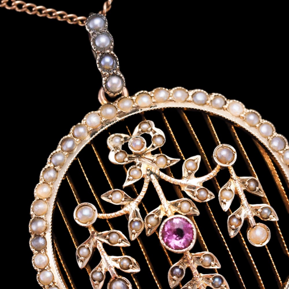 An Edwardian circular gold pendant featuring a halo of seed pearls framing an inner wire grid with a pearl-set floral garland and a central round pink tourmaline. View XV
